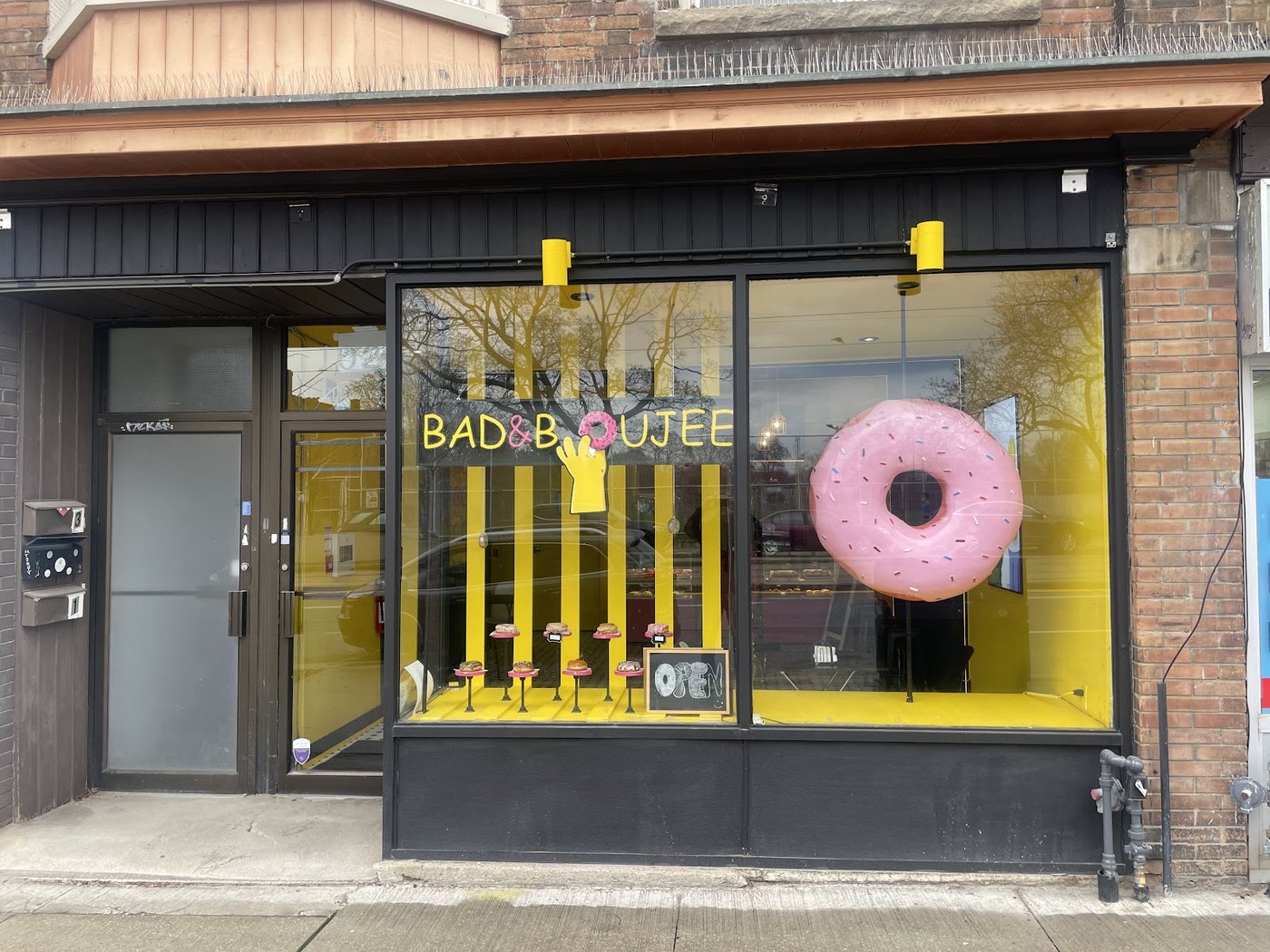 The Bad and Boujee shopfront on St Clair Avenue West: black frame, yellow interior, the wordmark and a large pink donut in the window